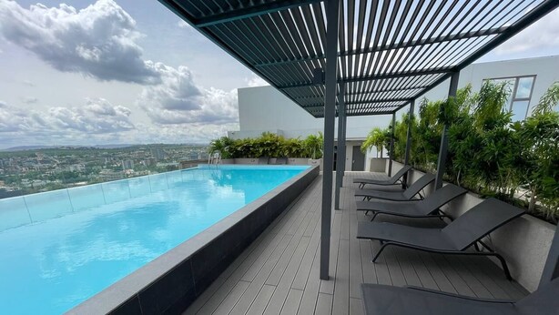 Piscina infinita en la azotea con terraza de madera y sillones, rodeada de vegetación y vistas a la ciudad.