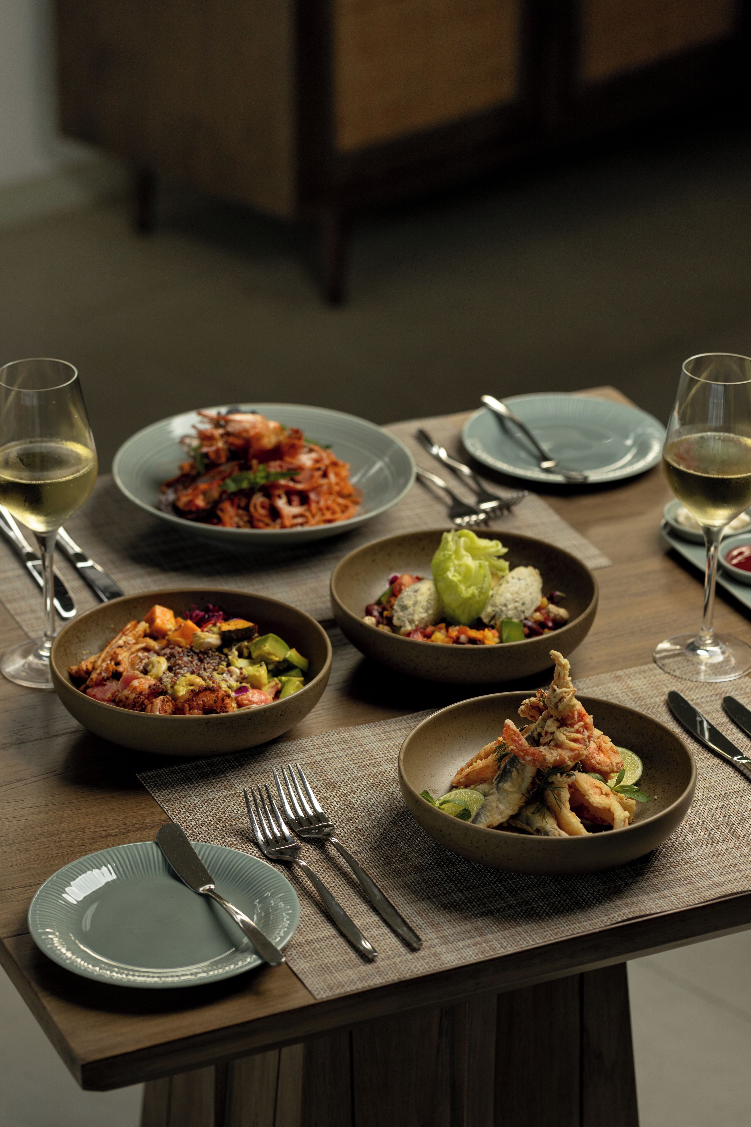 A dining table with multiple plates of food, cutlery, and glasses of white wine.