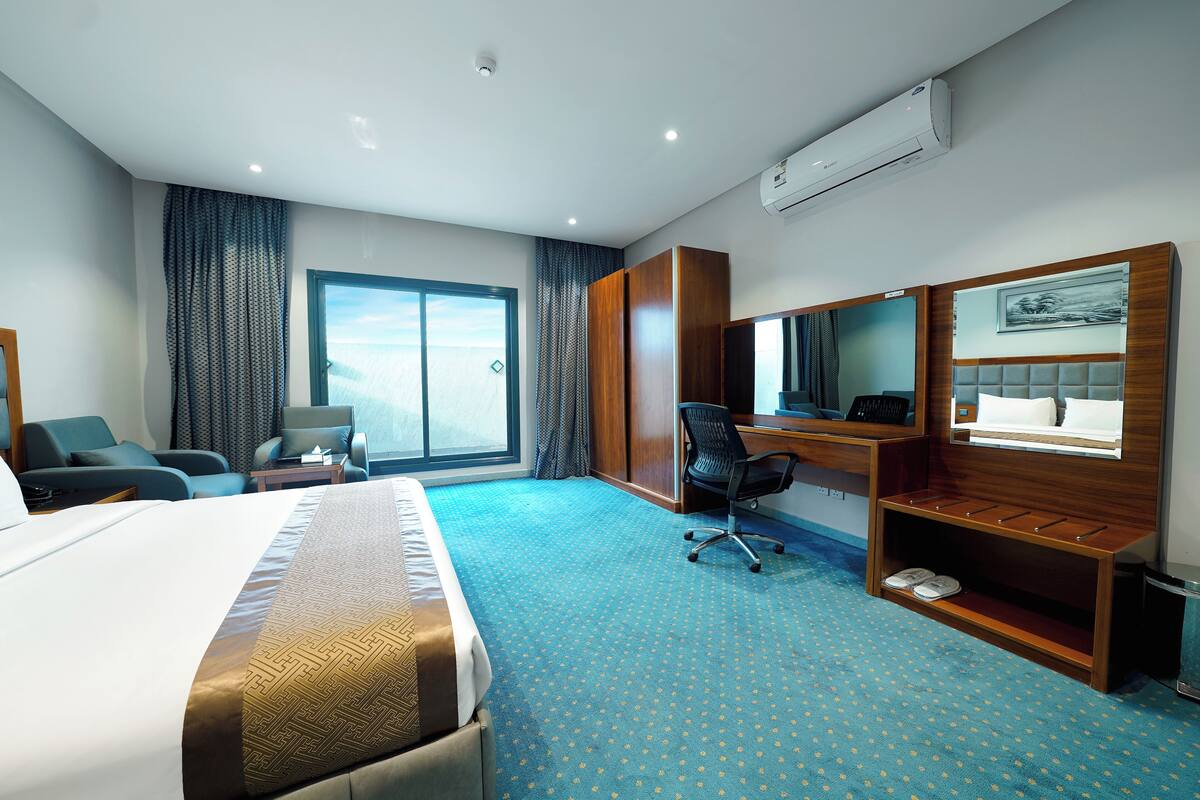 Een hotelkamer met een groot bed, een bureau met stoel, een klein tafeltje, een spiegel en een raam met gordijnen.