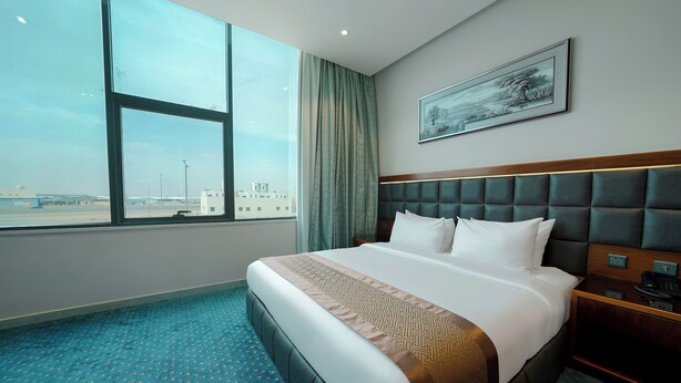 A hotel room with a large window, a bed with a headboard, a bedside table, and a painting on the wall. - Celine Home Serviced apartment -Tuwaiq  (Riyadh)