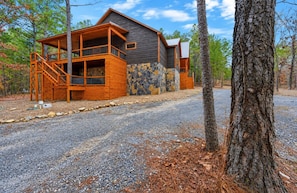 A cabin with a wooden deck and stone wall, surrounded by trees.
