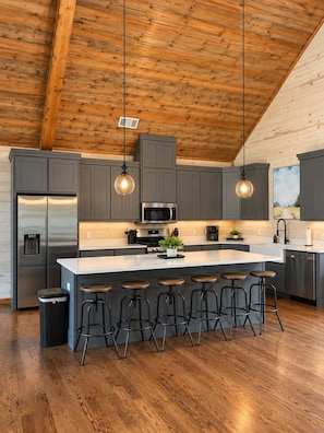 A modern kitchen with a central island, bar stools, stainless steel appliances, and pendant lighting.
