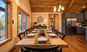 A dining area with a wooden table set for six, surrounded by chairs, with a kitchen in the background.
