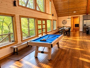 A wooden interior with a pool table, large windows, and a dining area.