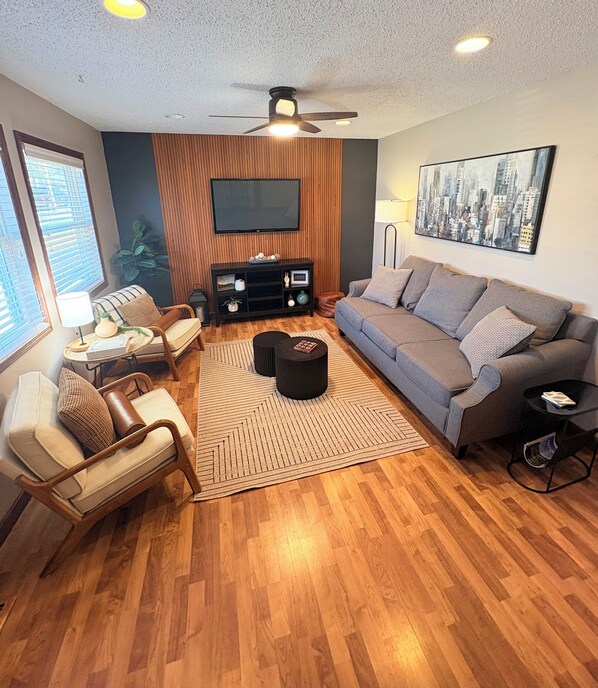 A modern living room with a grey sofa, wooden flooring, a ceiling fan, a flat-screen TV, and a cityscape painting.