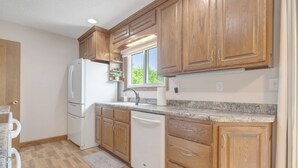 A kitchen with wooden cabinets, a white refrigerator, a dishwasher, and a window with a view of greenery. - The Modern Nest: Near Interstate, making it easy to get anywhere in Sioux Falls. (Sioux Falls)