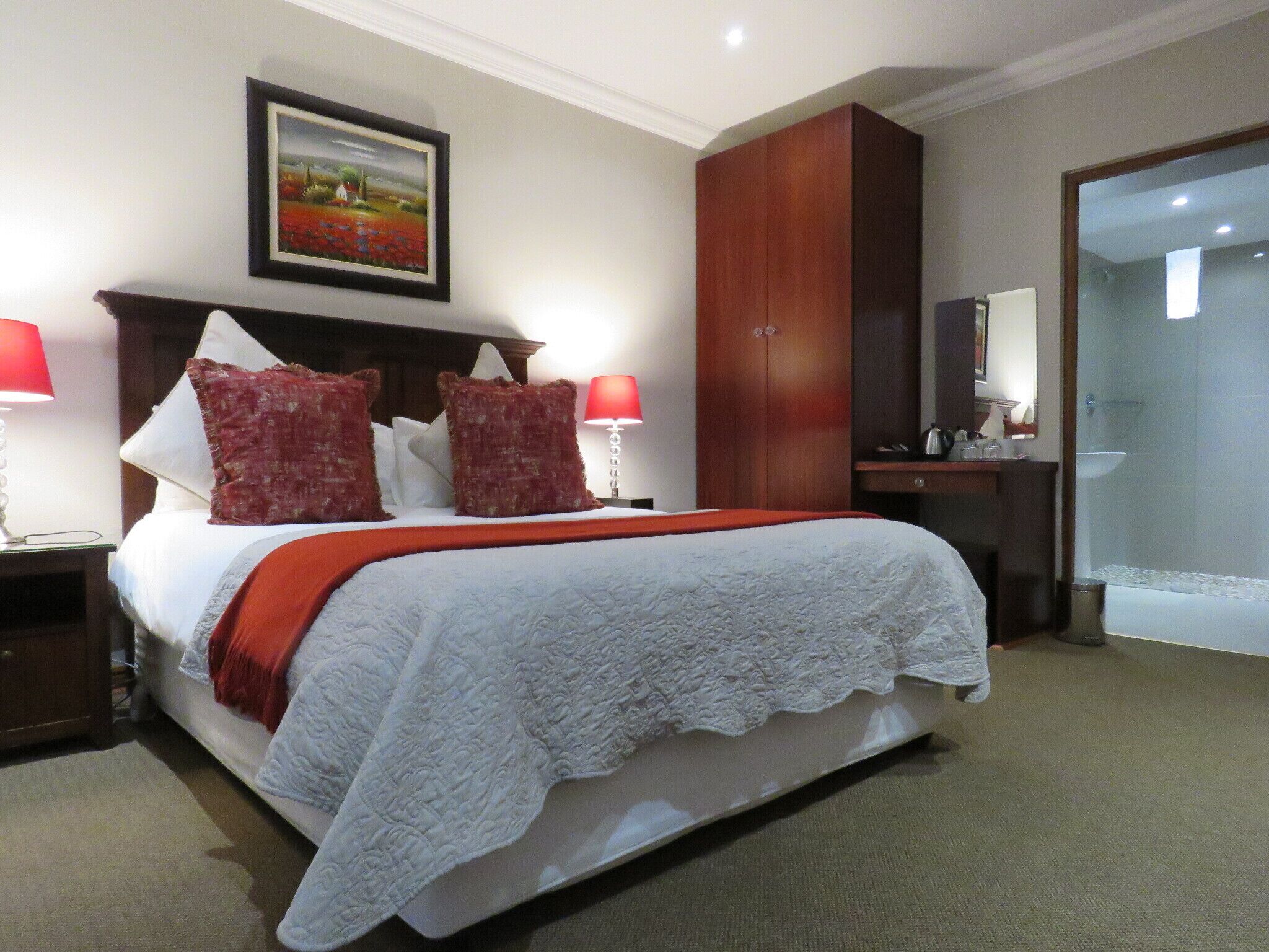 A hotel room with a large bed, two bedside lamps, a wooden wardrobe, a bathroom with a glass shower, and a framed picture on the wall.