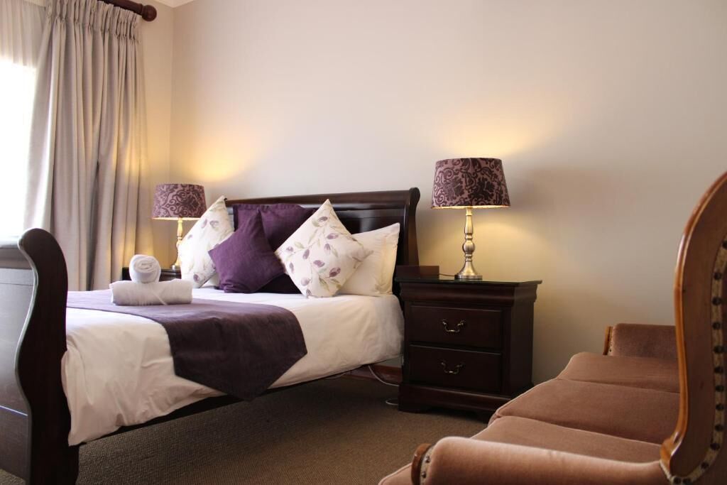 Una cama bien tendida con cabecera, dos lámparas de noche, una mesita de noche de madera y un sillón marrón.