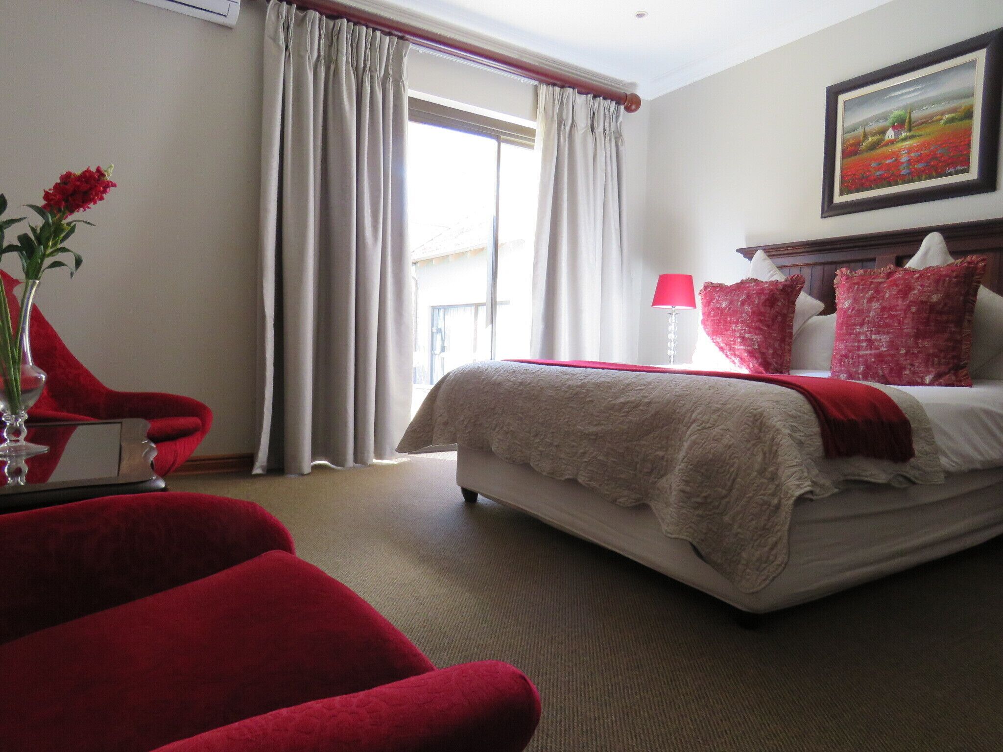 A hotel room with a bed, a red chair, a vase with flowers, and a painting on the wall.
