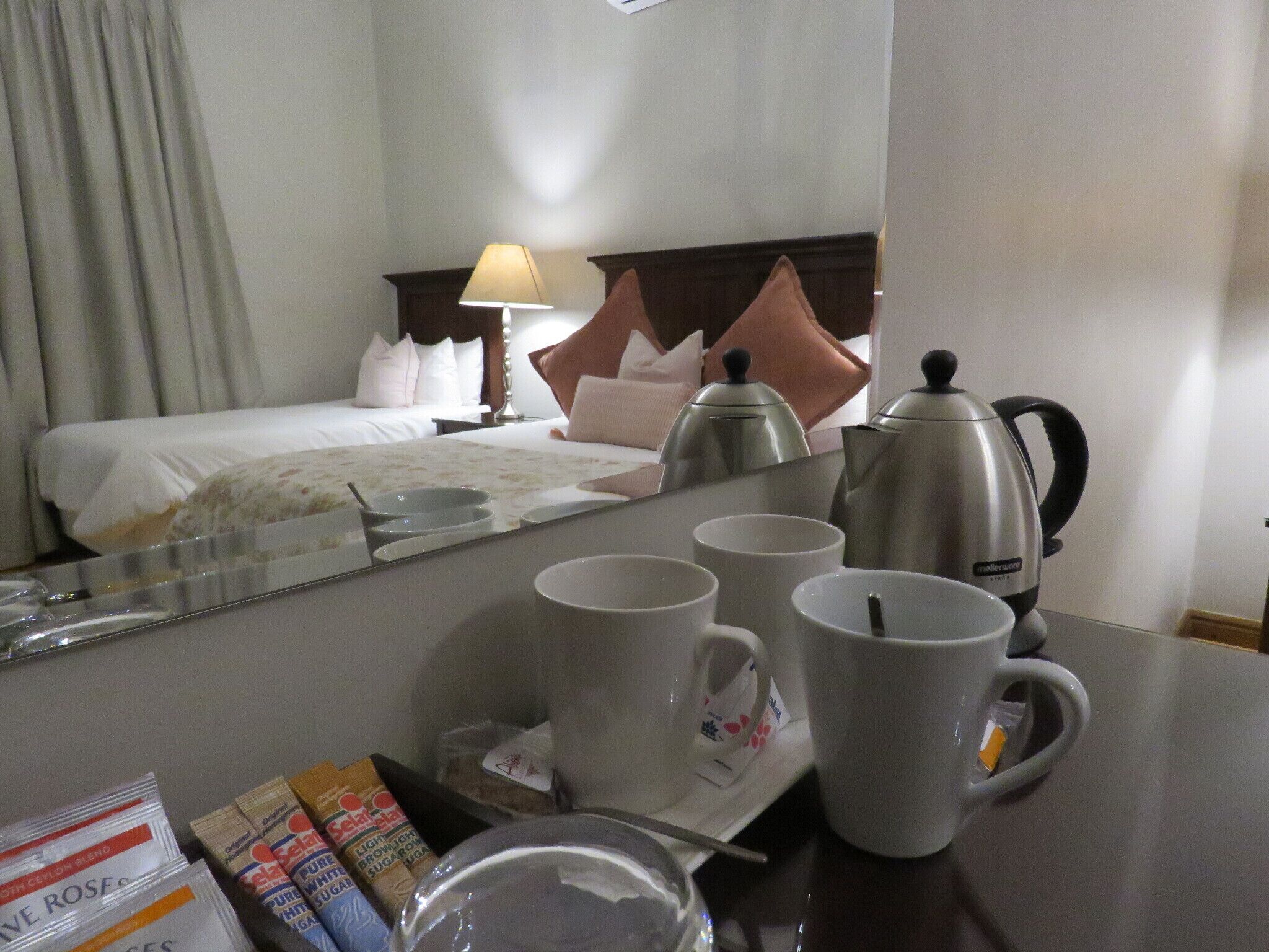 A hotel room with a bed, a nightstand, a lamp, a coffee maker, and a breakfast tray with cups and a glass.