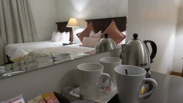 Quarto de hotel com cama, mesa de cabeceira, luminária, cafeteira e bandeja de café da manhã com xícaras e um copo.