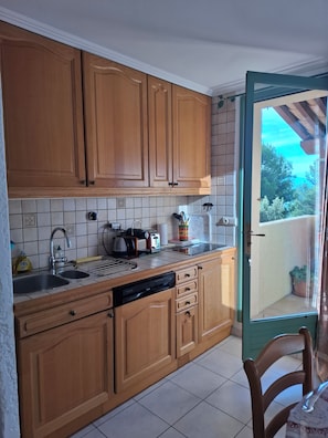 A kitchen with wooden cabinets, a dishwasher, and a window overlooking a balcony. - 8 Le Castellas (Sainte-Croix-du-Verdon)