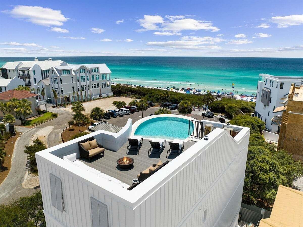 A rooftop pool area with lounge chairs and a fire pit, overlooking a beach and ocean.