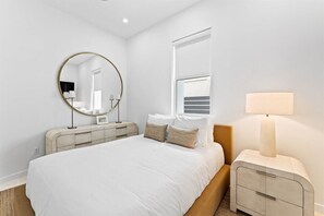 A modern bedroom with a large bed, a round mirror, a bedside lamp, and a window with blinds. - 30A | Rooftop Pool | Gulf Views | 2 Min To Beach! (Santa Rosa Beach)