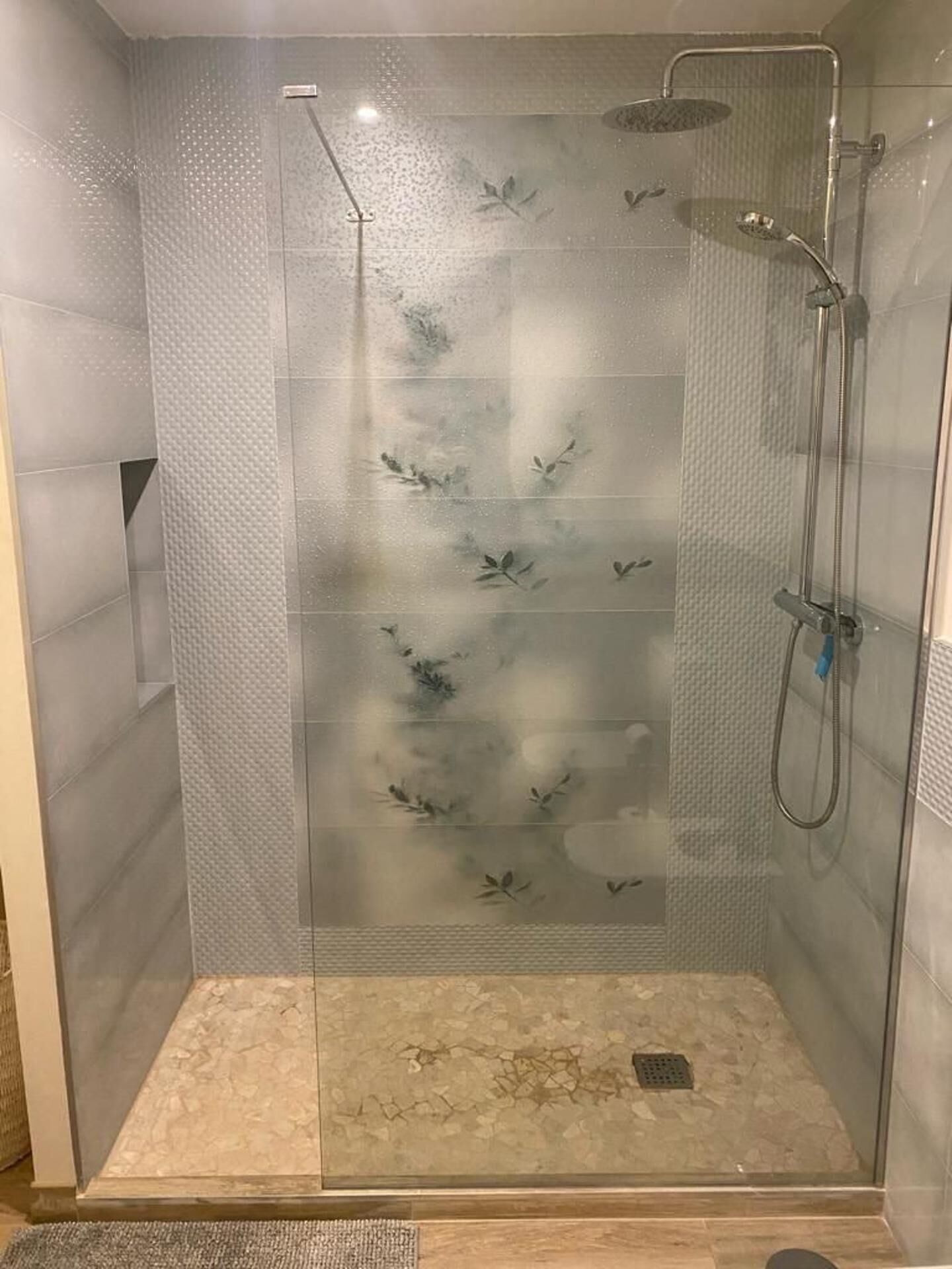 Eine Duschkabine mit Glaswänden, einer strukturierten Wand, einer Brause und einem Abfluss.