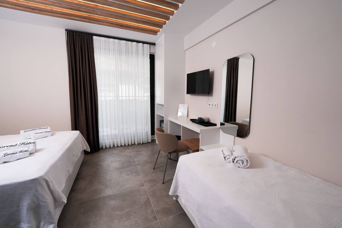 Ein Hotelzimmer mit zwei Betten, einem Schreibtisch, einem Sessel, einem Fernseher und einem großen Fenster mit Vorhängen.
