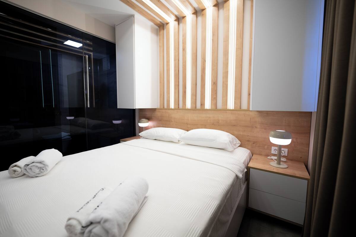Ein modernes Hotelzimmer mit einem großen Bett, einem hölzernen Kopfteil, einem Nachttisch mit Lampe und einer Glassturzwand.