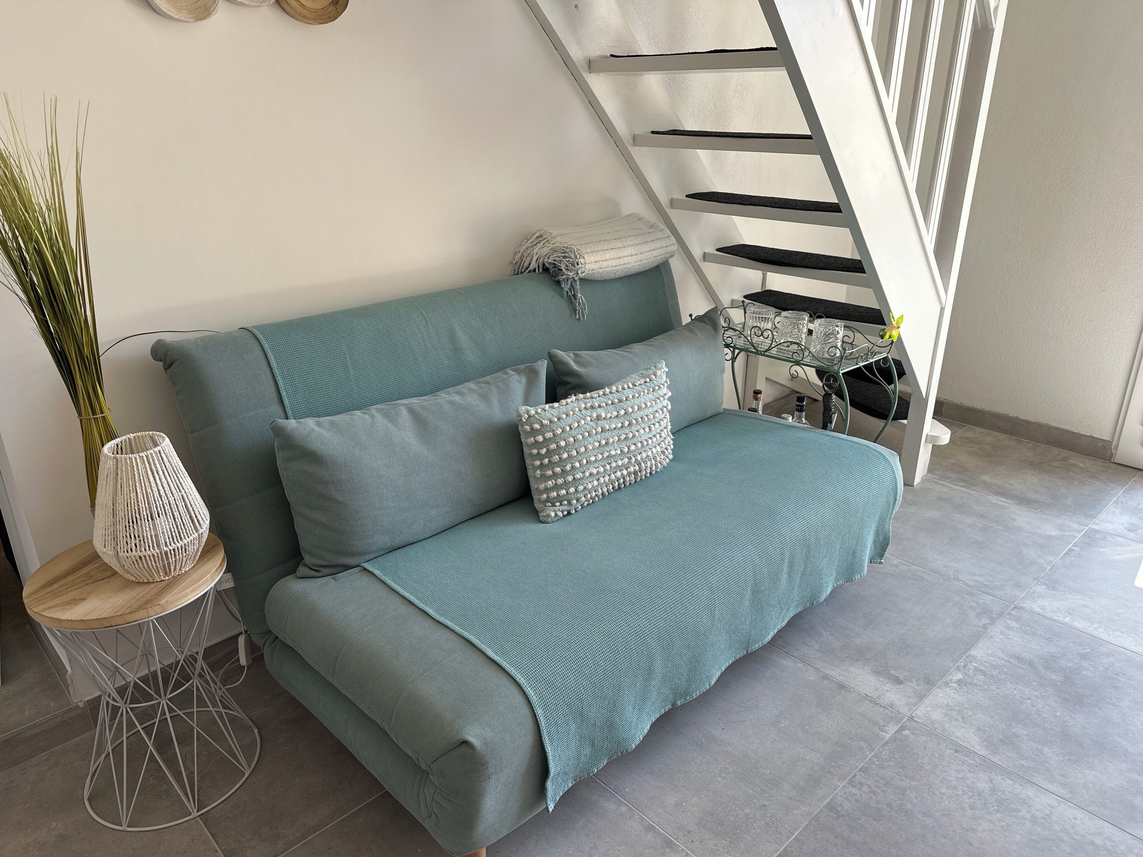 A teal sofa with a woven pouf and a small round table with a woven vase.