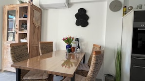 Een eethoek met een houten tafel, rieten stoelen, een kast met glazen deuren, een wandklok en een keuken met een koelkast.