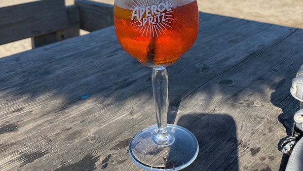 Een Aperol Spritz in een glas op een houten tafel, met een strand en boten op de achtergrond.