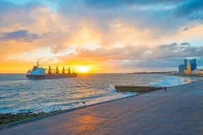 Ein Sonnenuntergang über einem Strand, im Vordergrund ein großes Frachtschiff und im Hintergrund die Skyline einer Stadt.