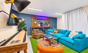 Ein Wohnzimmer mit einem blauen Sofa, einem Schachtisch, einem Sessel mit Leopardenmuster, einem Couchtisch und einem Neon-Schild mit der Aufschrift 'I LOVE THIS BAR'.