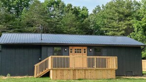 A single-story building with a dark exterior and a metal roof. It features a wooden deck with a staircase leading to a front door. - Black Butte - Lux Family Cabin With Games Firepit (Sevierville)