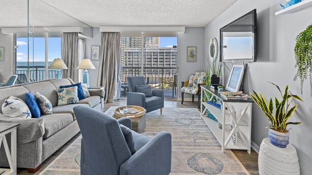 A modern living room with a grey sofa, armchairs, a coffee table, and a TV on a white stand. There is a large window with a view of buildings and a balcony. - S Hampton 1403 2 Br Oceanview in Kingston, Pool (Myrtle Beach)