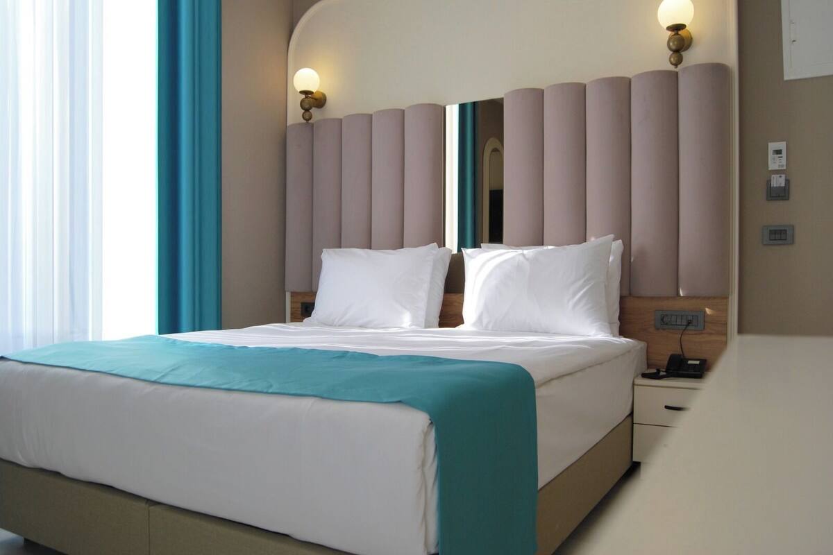 A hotel room with a bed, a nightstand, a wall-mounted lamp, and a window with curtains.
