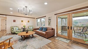 A spacious living room with a brown leather sofa, green patterned armchairs, a wooden coffee table, and a chandelier. A large sliding glass door leads to an outdoor deck with a view of the surrounding landscape.