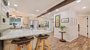 A modern kitchen with a large island, wooden stools, stainless steel appliances, and a view of the outside.