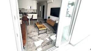 Una sala de estar moderna con un sofá, una mesa de centro y un televisor. Hay una zona de cocina con armarios y una mesa de comedor.