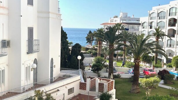 Un edificio blanco con balcones que dan a vistas del mar, palmeras y un cielo despejado.