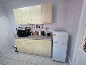 A compact kitchen with light wooden cabinets, a countertop with various appliances, and a white refrigerator.