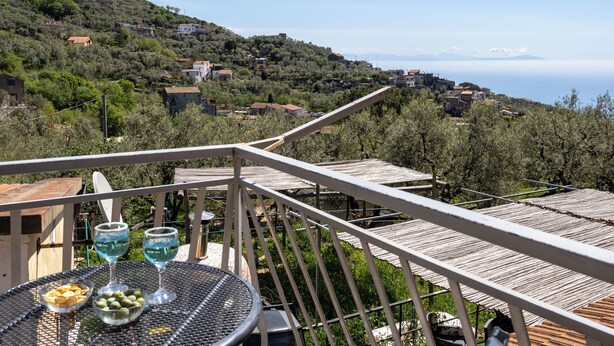 Una terrazza con un tavolo apparecchiato per due, affacciata su un pendio con edifici e vista sul mare.