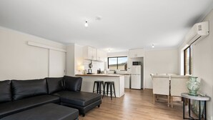 A modern living room with a black leather sofa, a dining area with a table and chairs, a kitchen with white cabinets, and a large window.