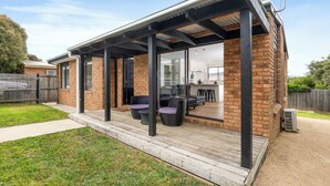 A covered outdoor patio with a brick wall, a wooden deck, and a glass door leading to an interior space with a kitchen and seating area.