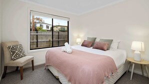 A bedroom with a bed, a chair, a nightstand, a lamp, and a window with a view of houses.