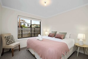 A bedroom with a bed, a chair, a nightstand, a lamp, and a window with a view of houses. - Jan Juc Living, Pet Friendly Coastal Escape for 6! (Jan Juc)
