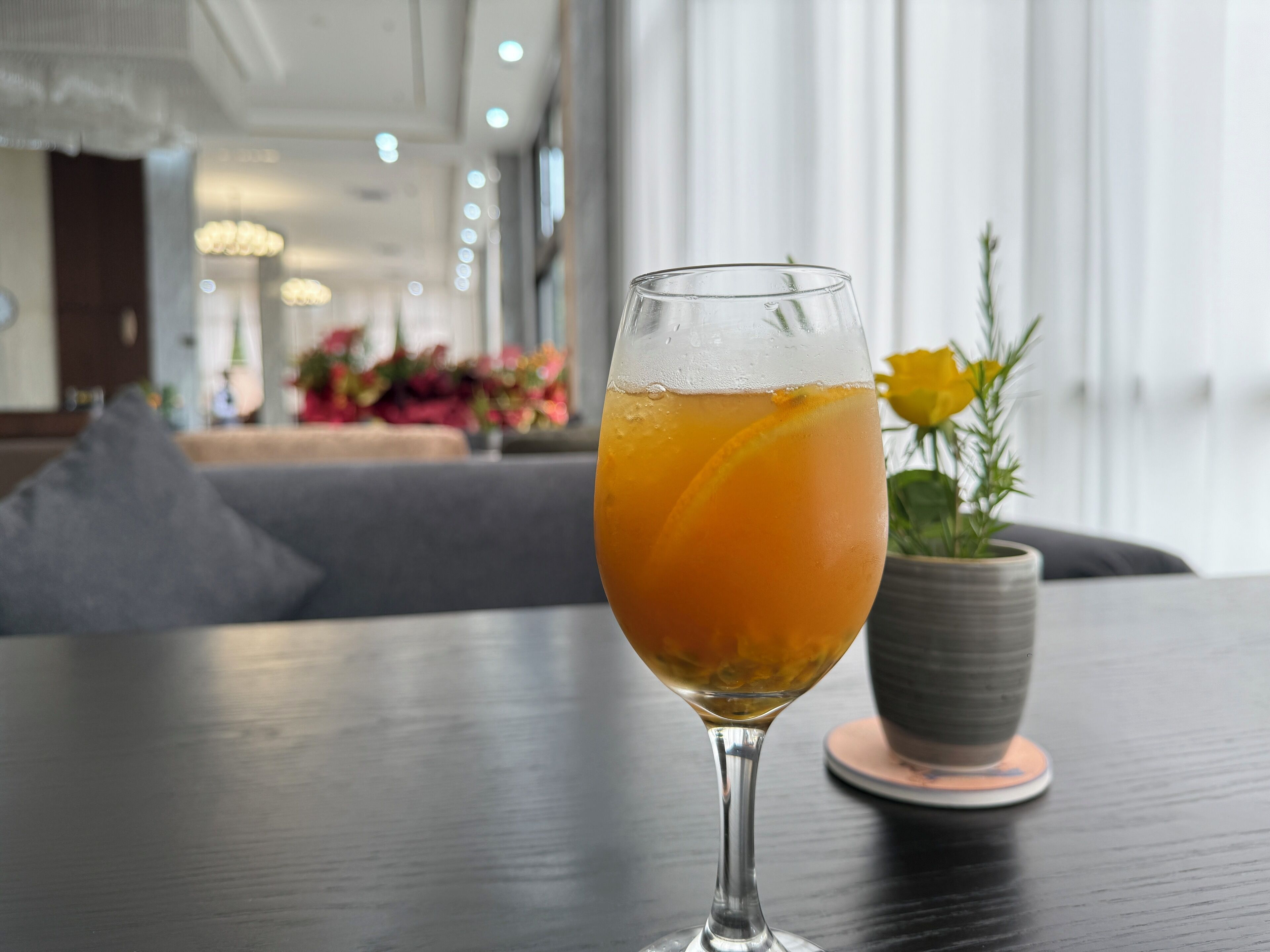 A glass of orange beverage on a table with a grey cushion and a potted plant in the background.