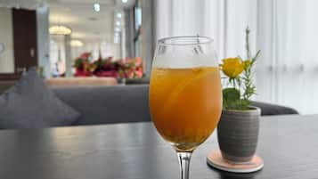 A glass of orange beverage on a table with a grey cushion and a potted plant in the background.