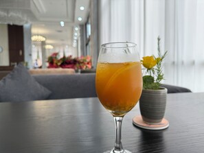 Un vaso de bebida de naranja sobre una mesa con un cojín gris y una planta en maceta al fondo.