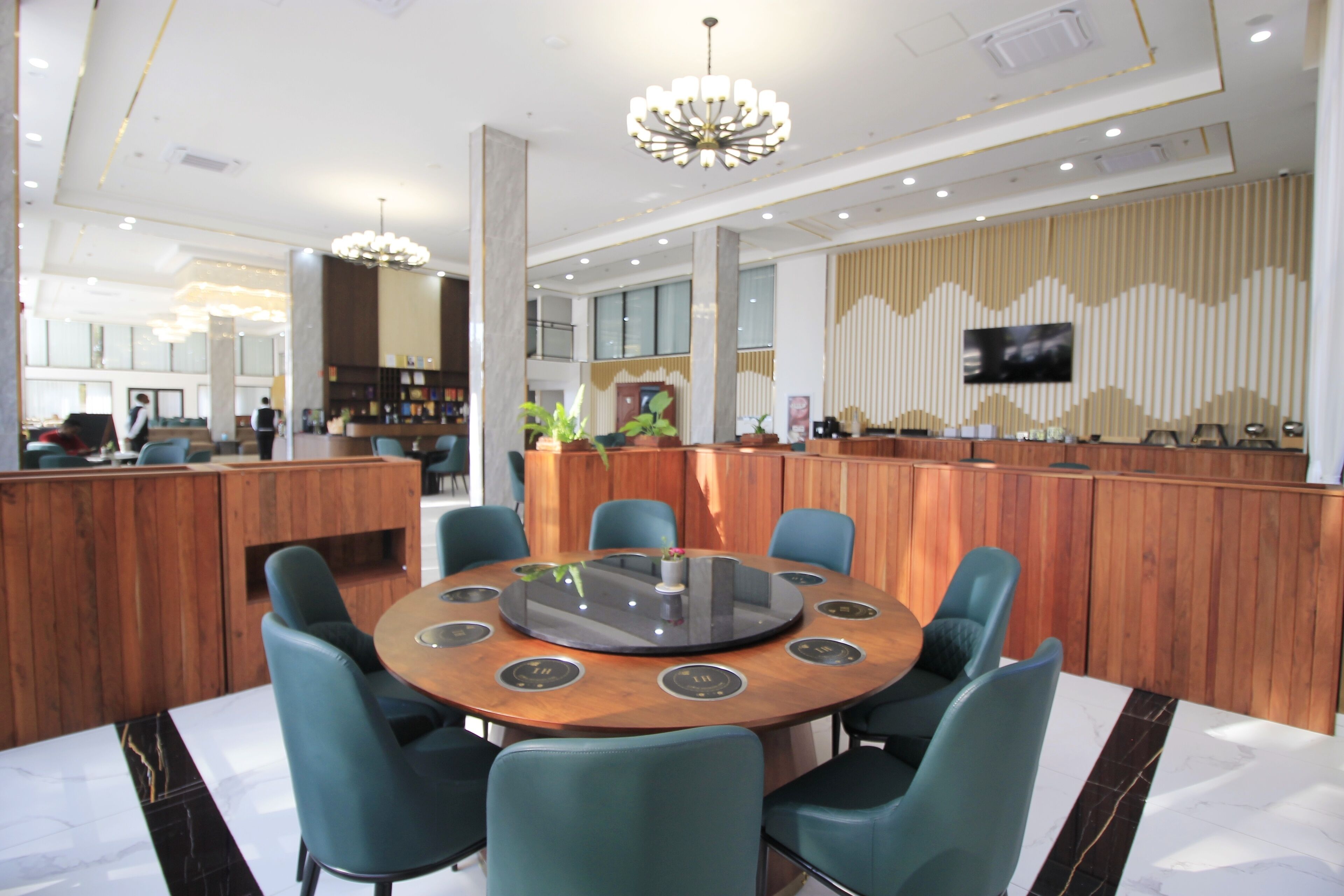 A modern hotel lobby with a round table, blue chairs, a wooden reception desk, and a large TV screen.