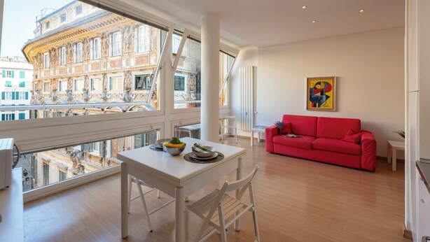 Een moderne woonkamer met een rode bank, een eettafel met stoelen en een groot raam met uitzicht op een stadsbeeld.