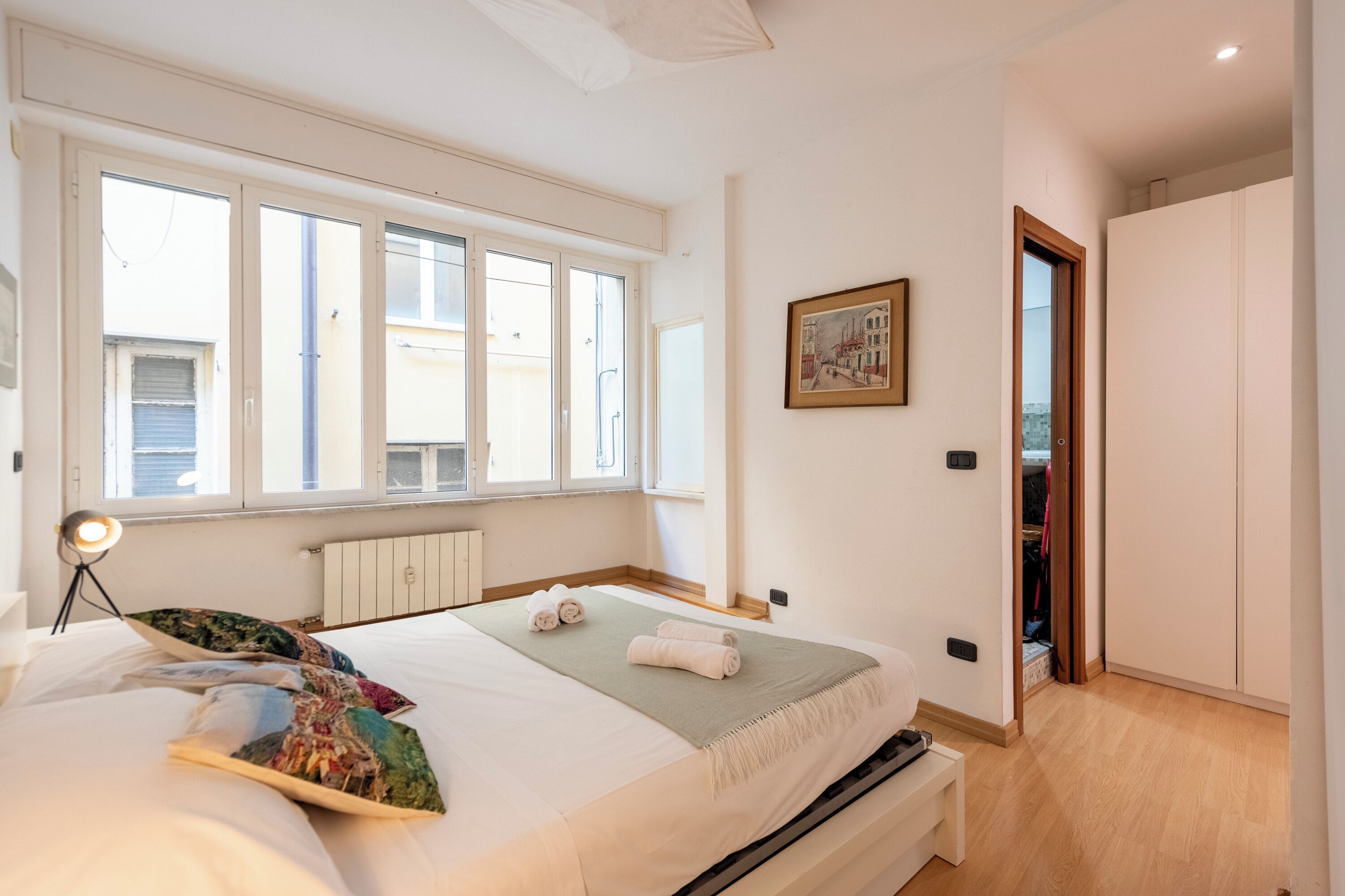 A bedroom with a bed, a bedside lamp, a window, a painting, and a wardrobe.