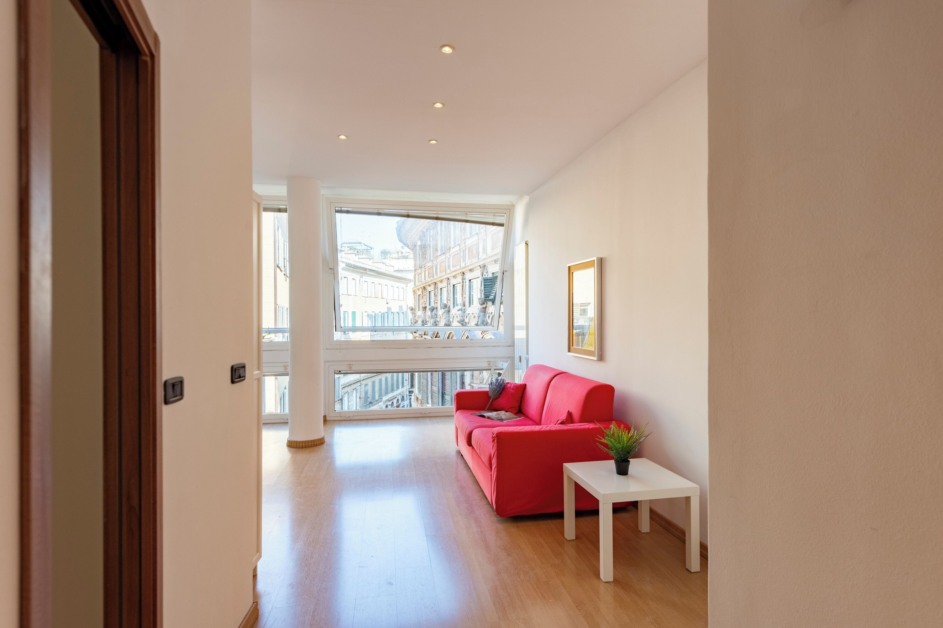 Ein geräumiger Wohnbereich mit einem roten Sofa, einem kleinen weißen Tisch und einem Blumentopf. Ein großes Fenster bietet Blick auf die Stadtlandschaft.