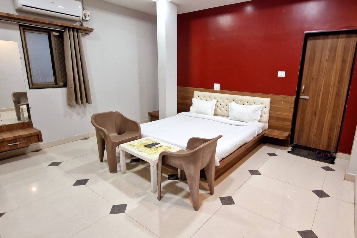A hotel room with a bed, a table, two chairs, a wooden wardrobe, and a red accent wall.