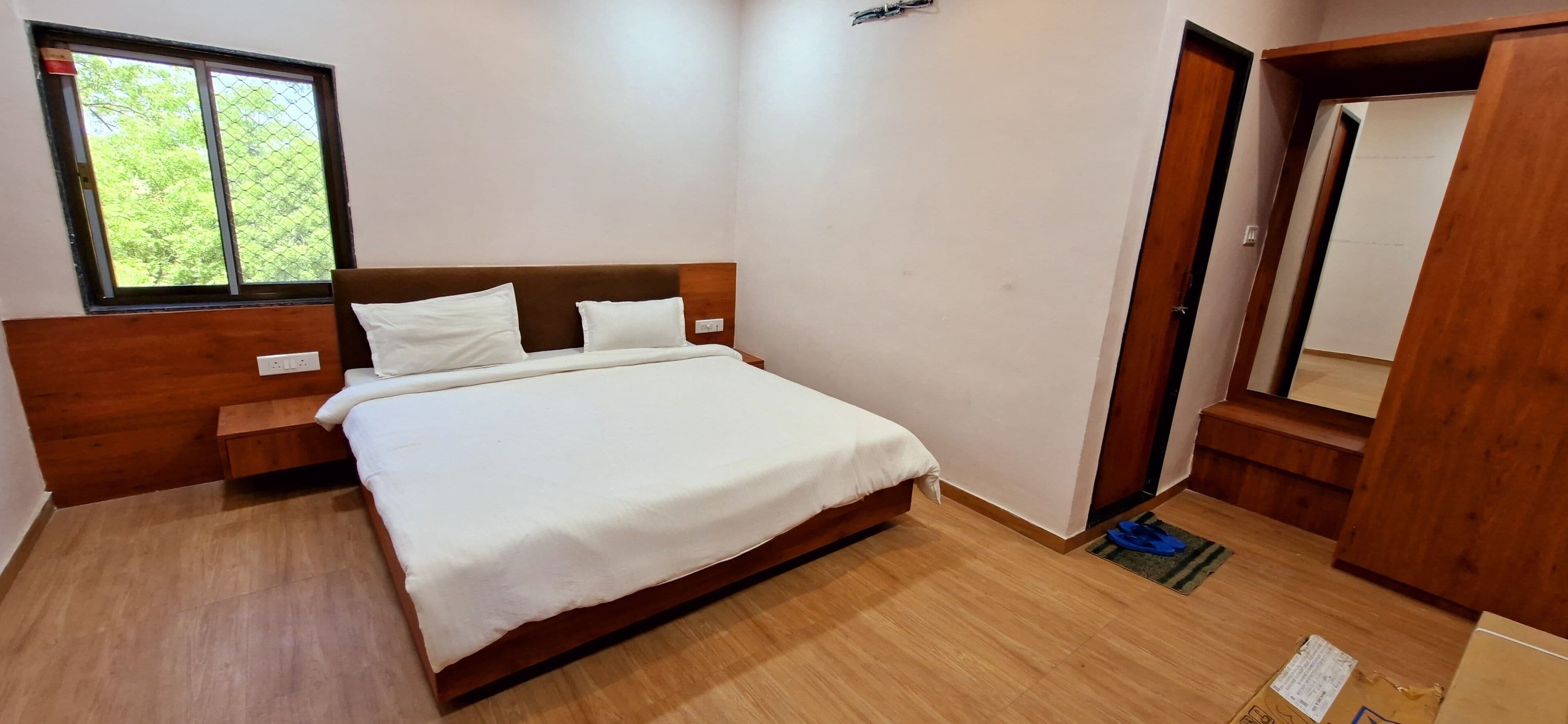 A neatly arranged bedroom with a wooden headboard, a large bed with white linens, a window with a view of greenery, and a wooden door.