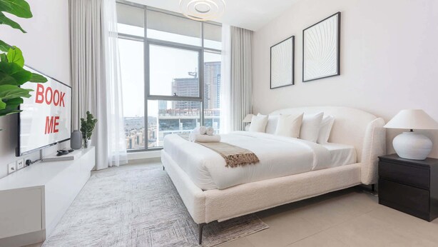 A modern hotel room with a large bed, a TV on the left, and a city view through the window. - StayShort - Stylish Modern Studio in Living Garden 2 (JVC)