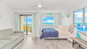 Une chambre avec un lit, un bureau, une chaise, un ventilateur de plafond, une fenêtre donnant sur la mer et un balcon avec une chaise.